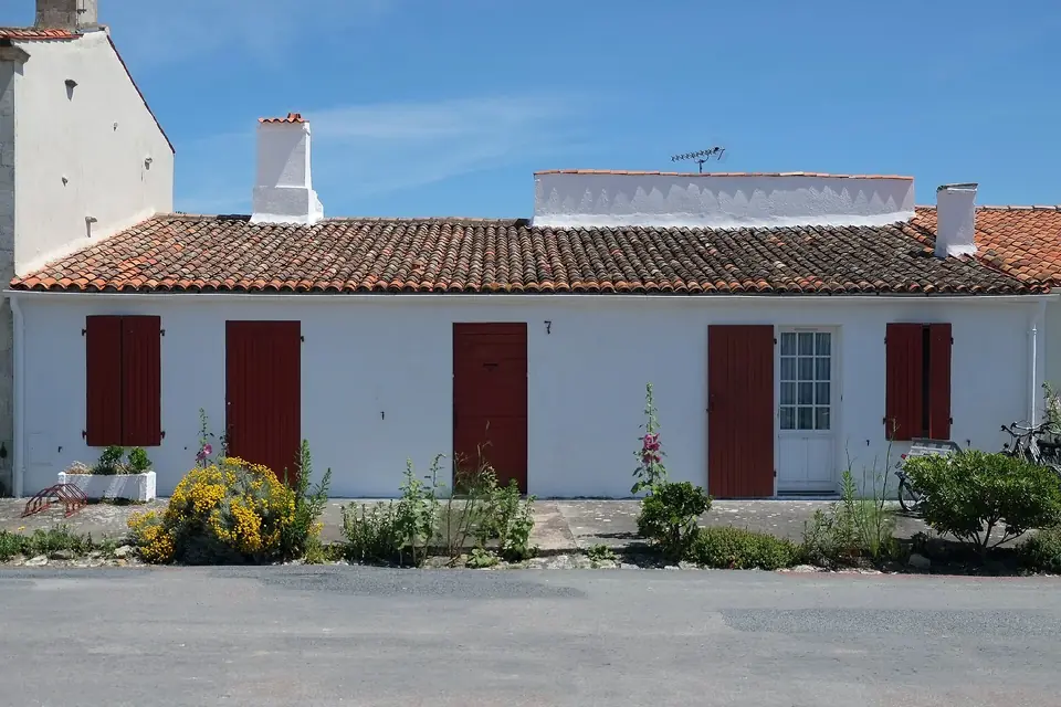 Maison basse de l'île d'Aix aux volets battants rouges fermés, façade blanchie et massifs fleuris le long du trottoir.